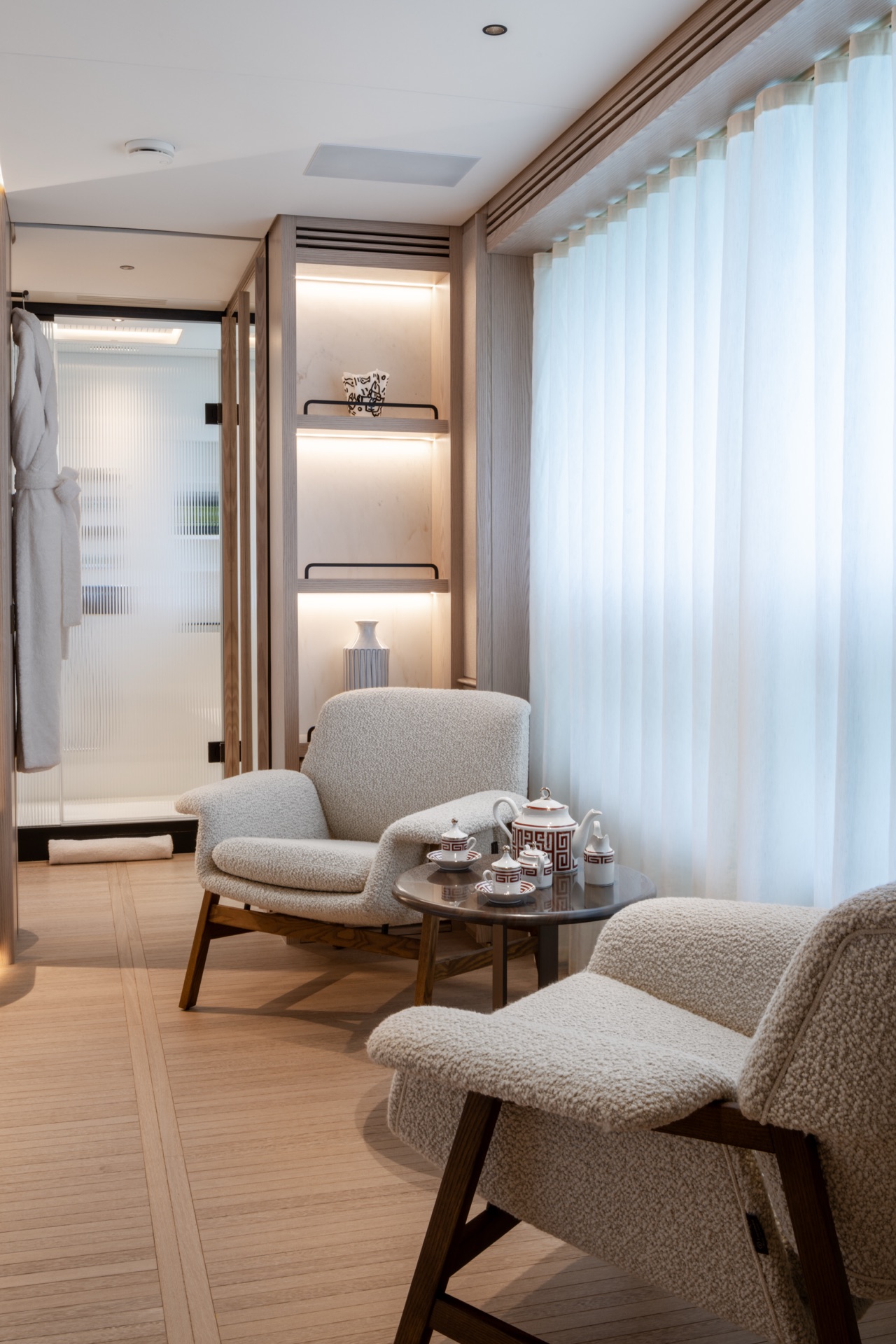 Two bouclé armchairs and a tea set by the window, with a lit display niche and the bathroom beyond