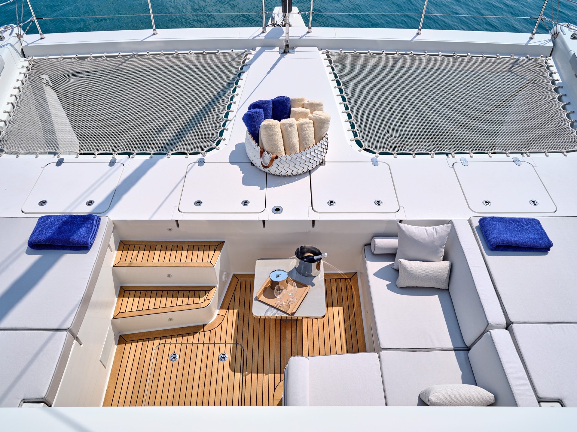 Overhead view of the catamaran's foredeck: trampoline netting, teak steps and a basket of rolled towels