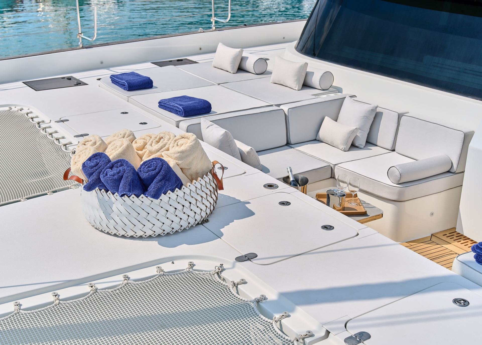 Catamaran foredeck with white sun pads, cushions and a woven basket of rolled navy and white towels