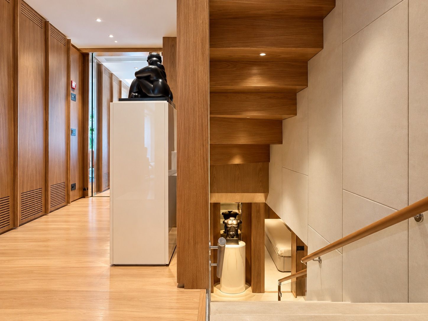 Lower deck corridor in pale oak and light stone, with a bronze sculpture on a white plinth beside the staircase