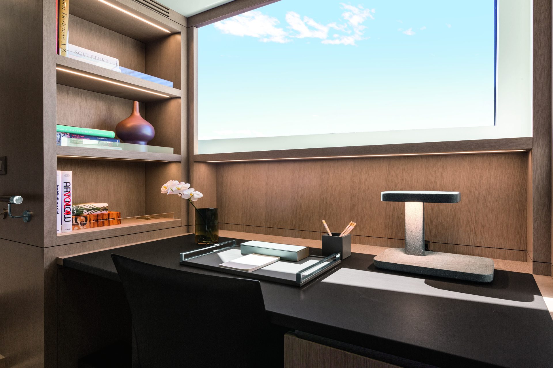 Desk in dark lacquer below a window, with a brass lamp, books and an orchid